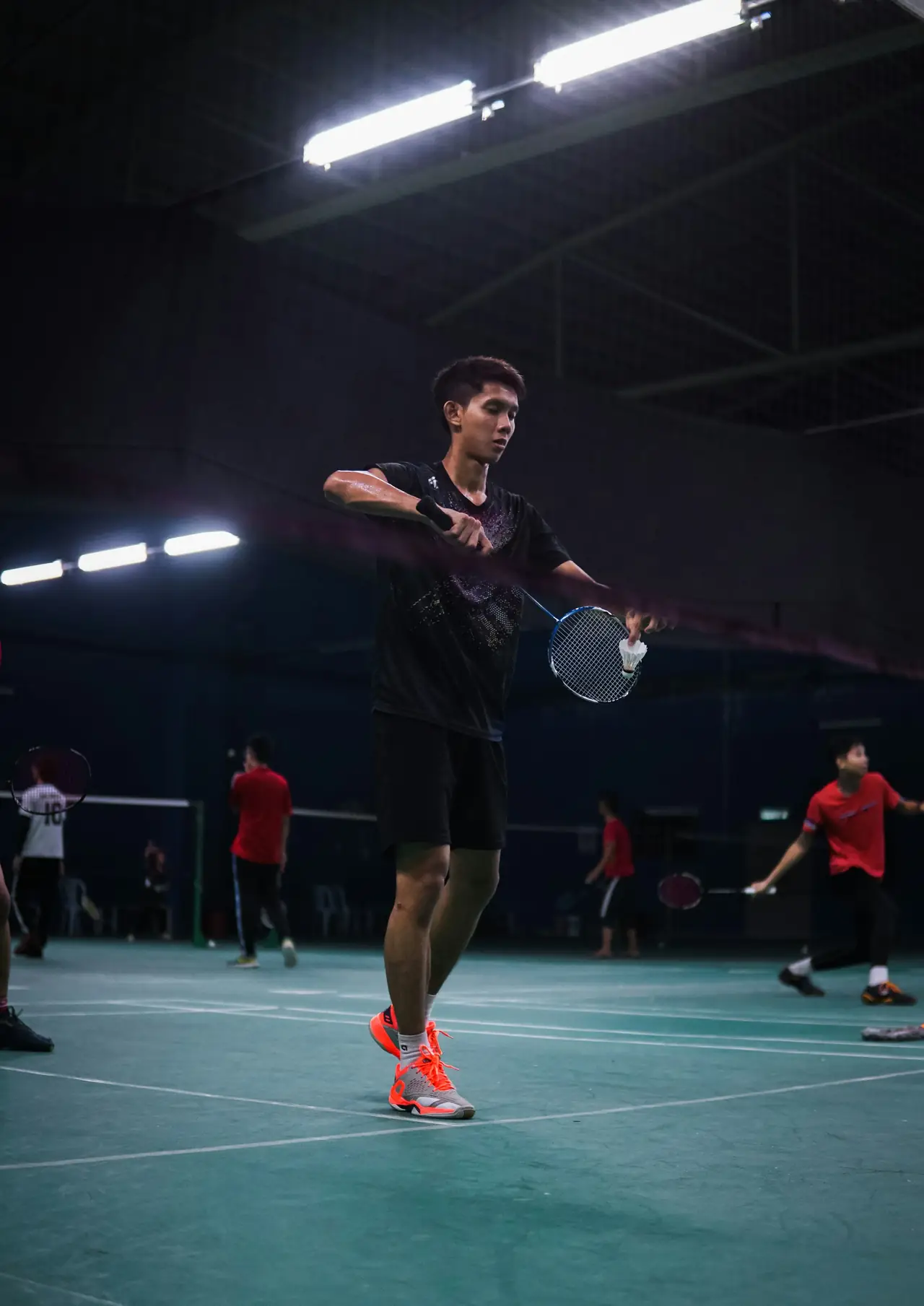 Players rallying on an indoor court under floodlights, with other matches going on behind.