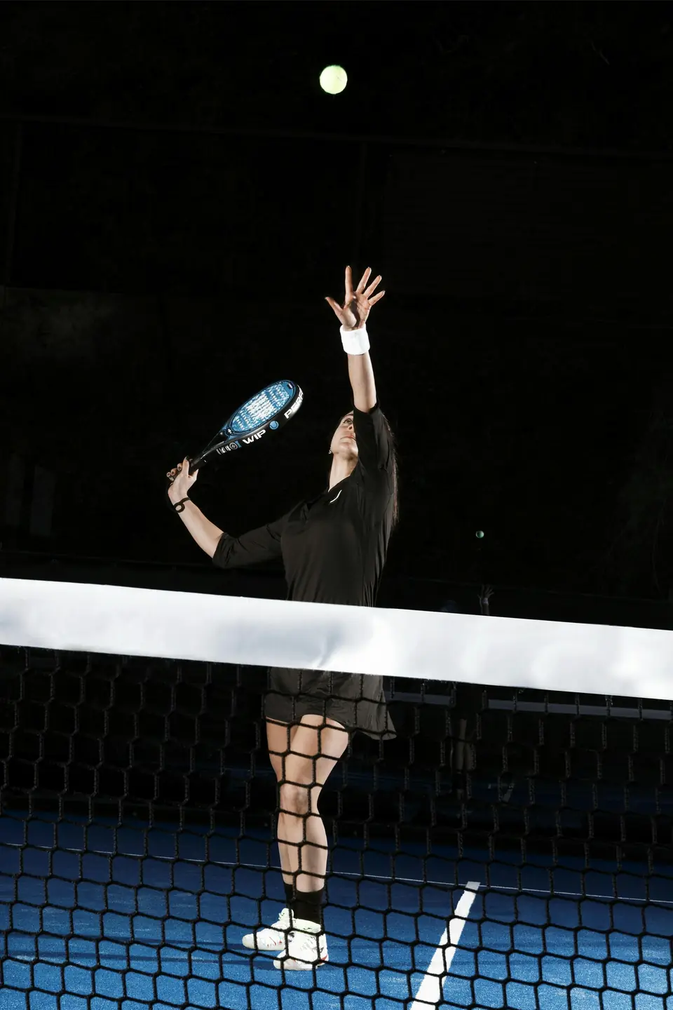 A player reaching up to serve, lit against a dark court.